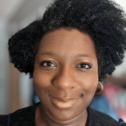 Abiola Agbayewa with black and grey natural Afro hair, wearing gold hoop earrings against a blurred background.