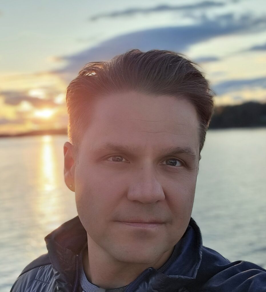Jeff Stecyk with short brown hair, wearing long-sleeved shirt with collar up, standing in front of a a sunset over a body of water.